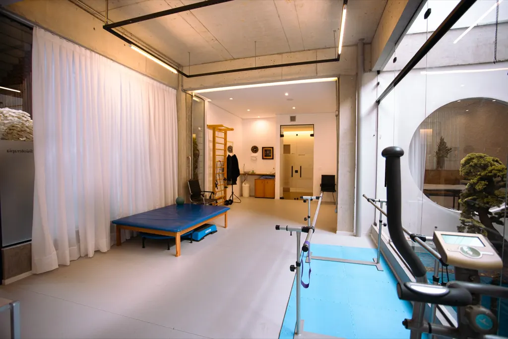 Sala de reabilitação da clínica com tatame azul, espaldar e barras paralelas.