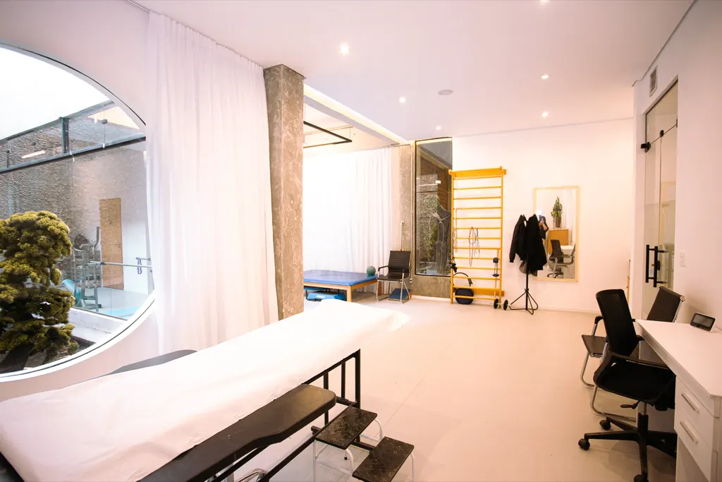 Sala de atendimento da clínica com janela redonda, maca e mesa.