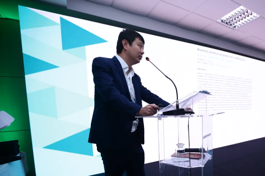 Dr. Marcus Yu Bin Pai, de paletó escuro, fala ao microfone em um púlpito de acrílico, diante de um telão com o slide “Western medical acupuncture: a definition”.
