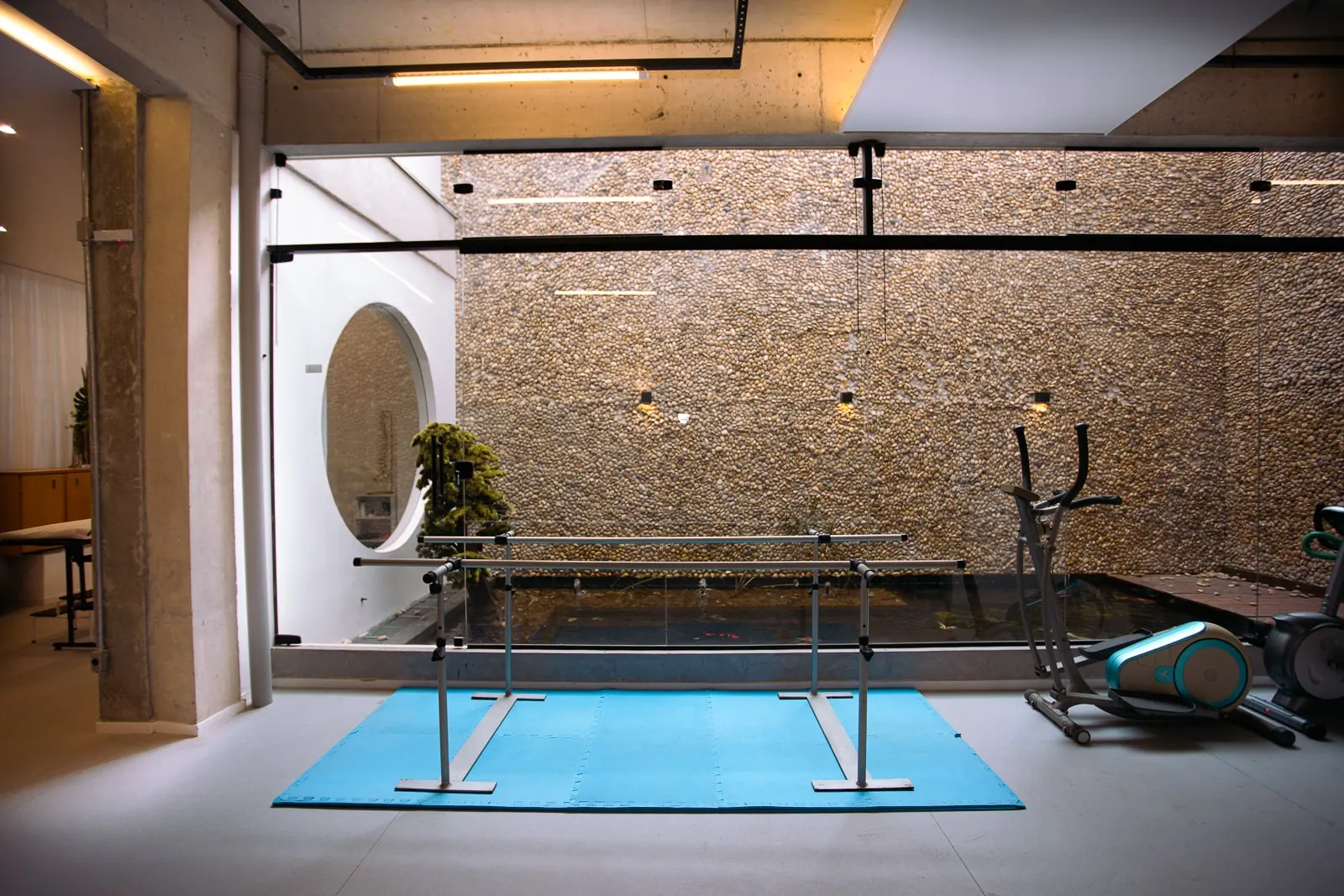Sala de reabilitação da clínica com parede de pedra, barras paralelas e tatame azul.