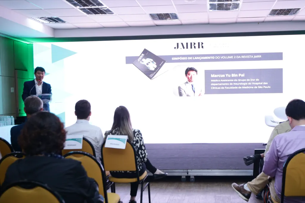 Sala de evento vista do fundo: Dr. Marcus Yu Bin Pai fala ao púlpito, à esquerda, diante da plateia sentada e de um telão com o seu nome.