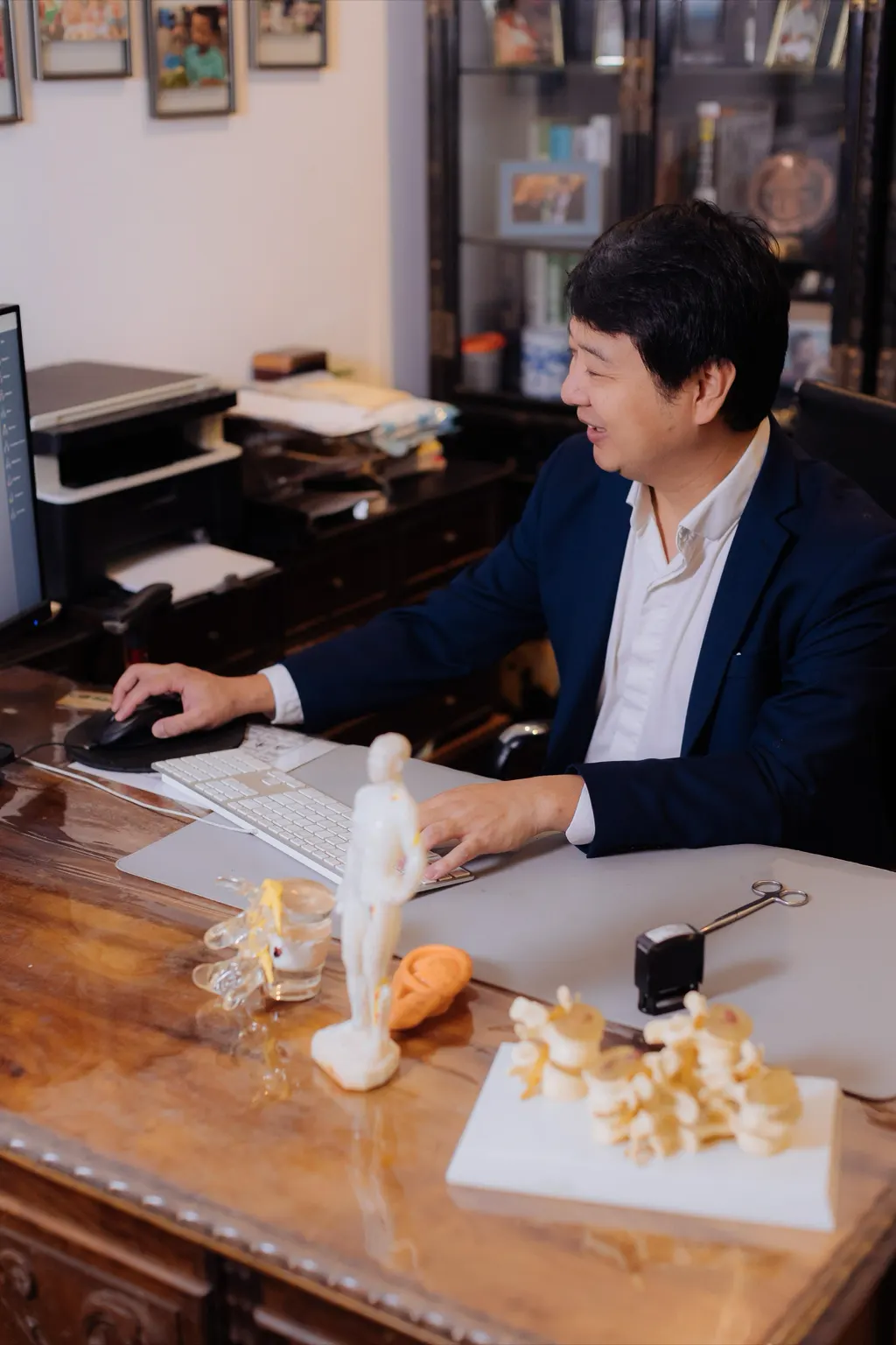 Dr. Marcus Yu Bin Pai, de perfil, trabalha à mesa do consultório, com modelos anatômicos em primeiro plano.