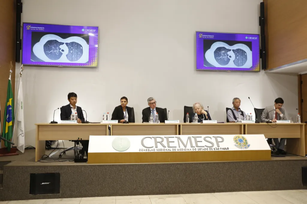 Mesa de evento no CREMESP vista da plateia, com seis participantes e a placa do CREMESP à frente.