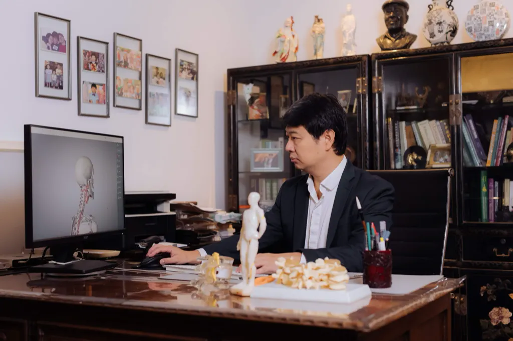Dr. Marcus Yu Bin Pai trabalhando à mesa do consultório, com modelos anatômicos e estante de livros.