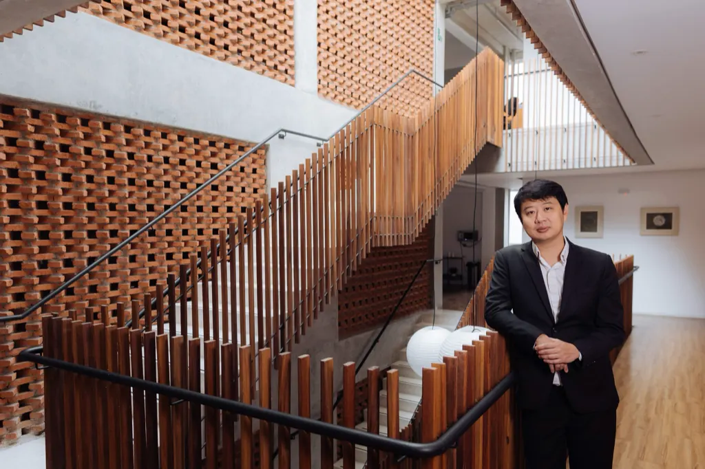 Dr. Marcus Yu Bin Pai em pé junto ao corrimão, diante da escada de ripas de madeira e da parede de tijolos vazados.
